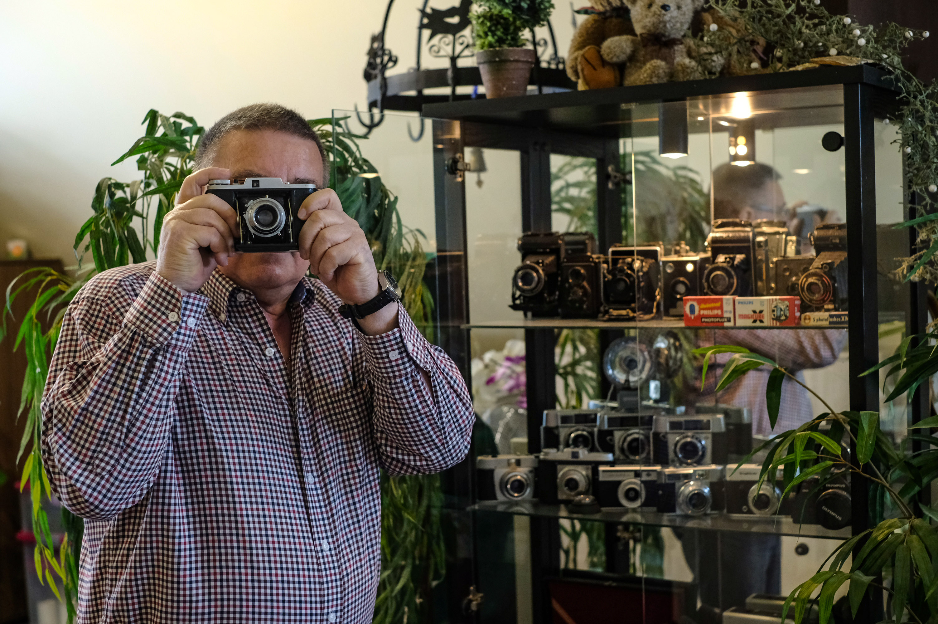 Mann hält sich alte Kamera vor das Gesicht und steht neben Schrank mit vielen alten Kameras.